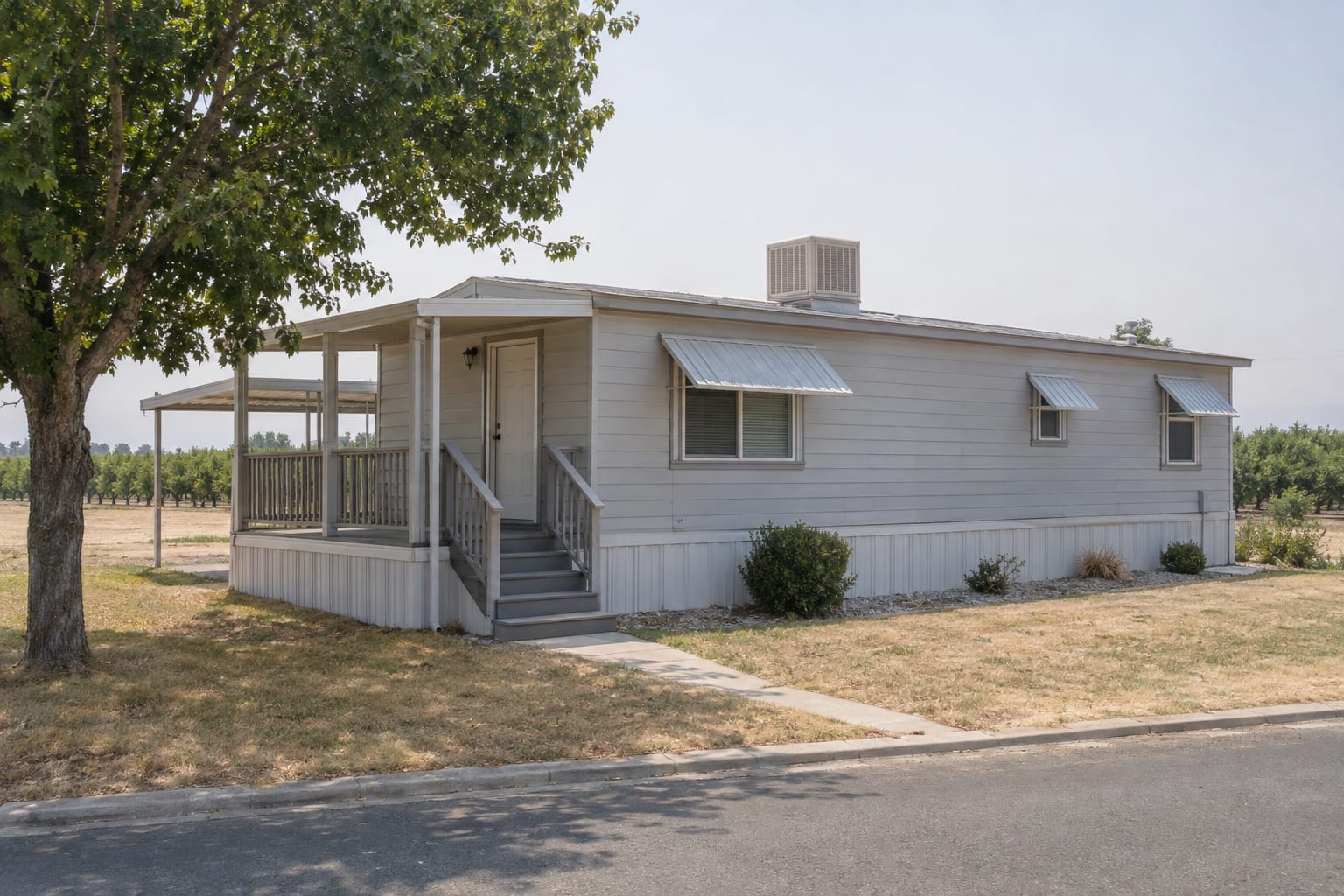 A single-wide manufactured home with a rooftop evaporative cooler and metal awnings shading its windows, in hazy summer light.