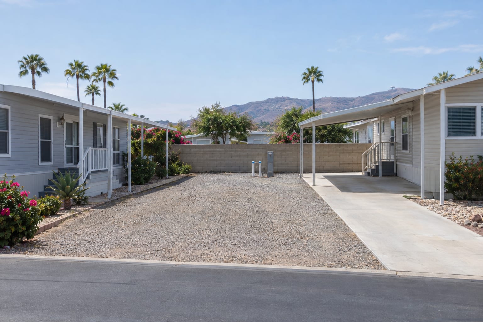 An empty gravel homesite with a concrete driveway strip and a utility pedestal, between two manufactured homes in a community