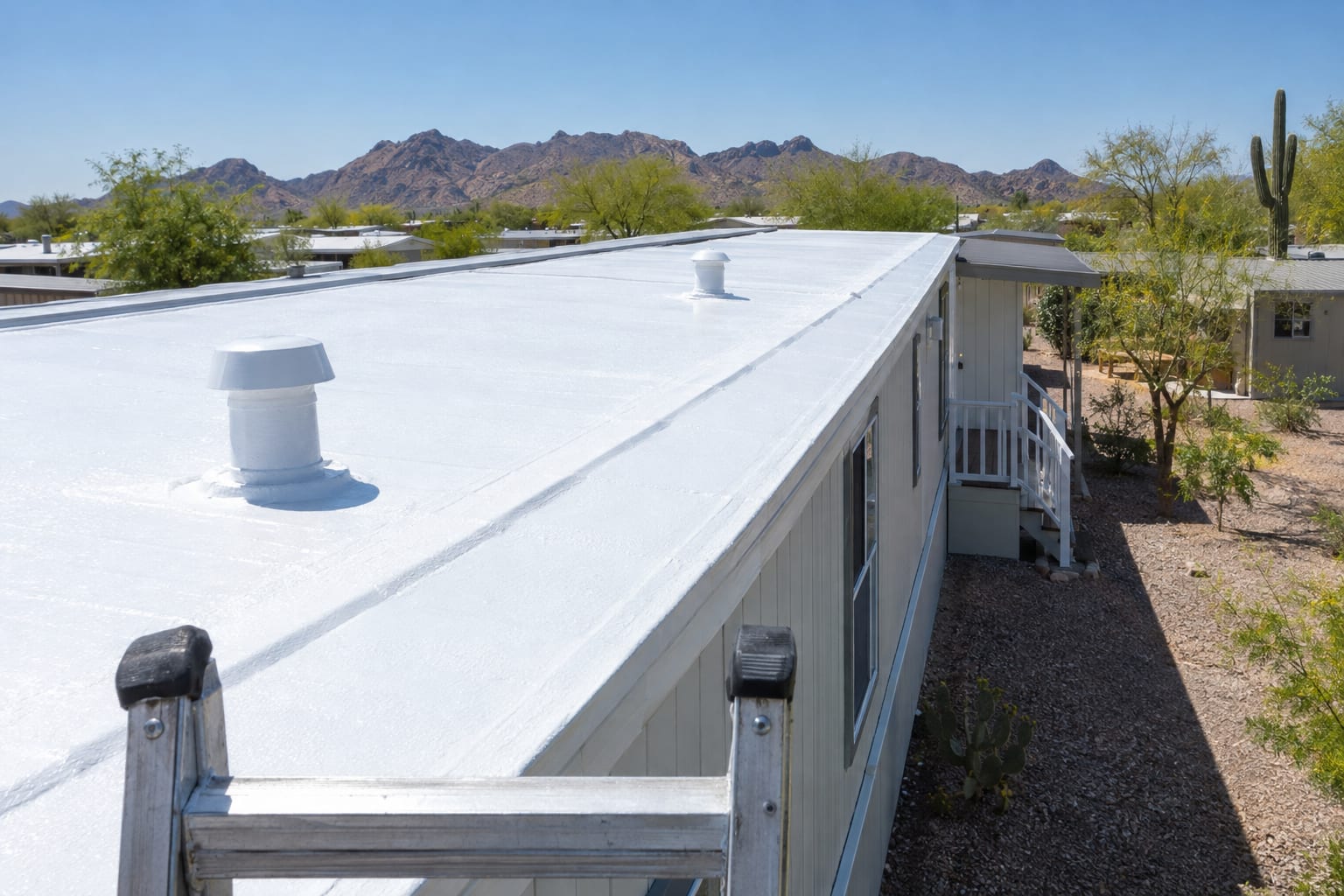 The low-slope metal roof of a single-wide manufactured home coated bright white, seen from the top of a ladder at the eave, with desert hills in the distance.