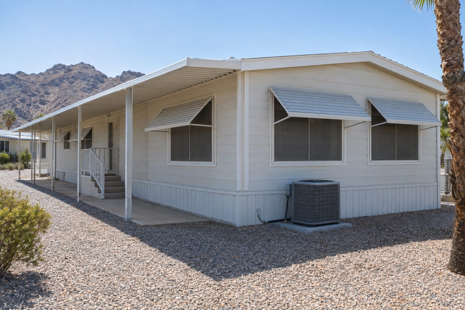 The sunny side of a manufactured home in the desert, with metal awnings over the windows, a shaded patio cover and an air-conditioning unit on a concrete pad